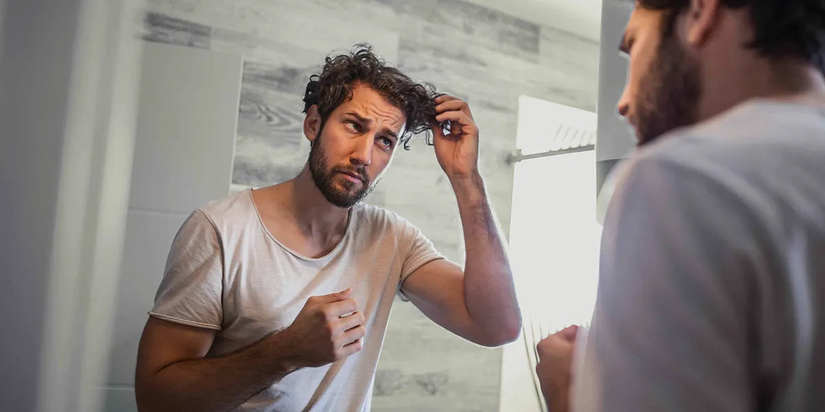 Young man looking in mirror touching hair wondering about causes of hair loss.
